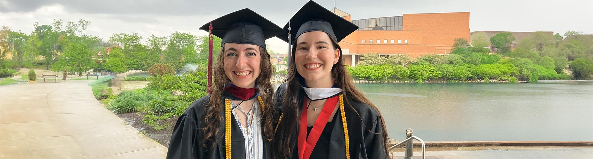 Emily Vest and Brianna De Man celebrate their graduation from Cedarville University in their caps and gowns with Cedar Lake and the Dixon Ministry Center in the background